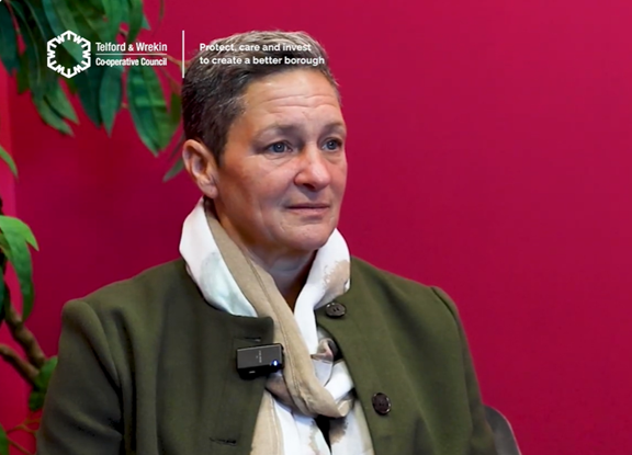 A person wearing a dark green coat and a light-coloured scarf is seated indoors against a red wall. A small microphone is clipped to the coat. There is a green leafy plant partially visible on the left side of the image. White text in the top left corner reads “Telford & Wrekin Cooperative Council,” and additional text nearby says, “Protect, care and invest to create a better borough.”